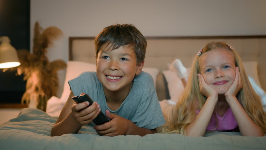Happy smiling little caucasian children boy girl brother sister watching TV together lying on bed in bedroom kids sibling watch cartoon hold remote control having fun laughing at home enjoy television | Shutterstock HD Video #1109212893