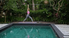 Mid adult woman standing in yoga pose by swimming pool on sunny day at resort. High quality 4k footage
