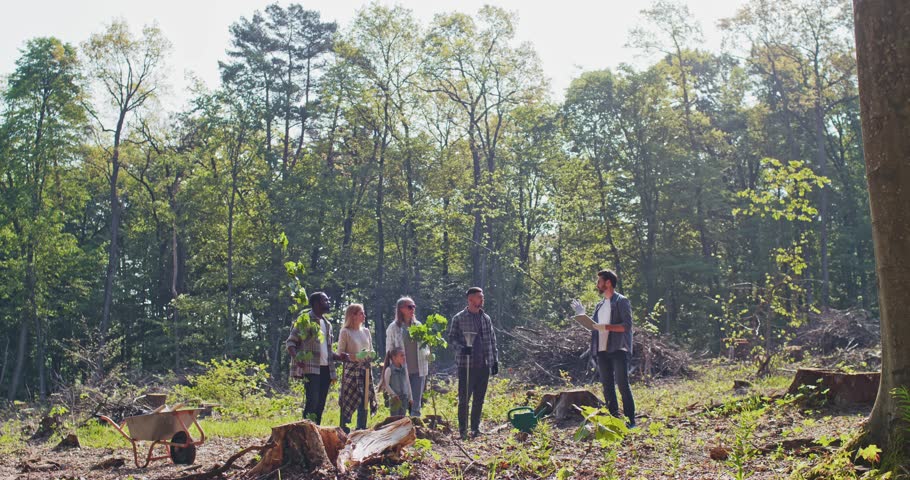 Multi-aged multicultural group of caring people volunteers planting forest listening to instructor standing in beautiful forest. Enthusiastic people providing forestation helping nature.