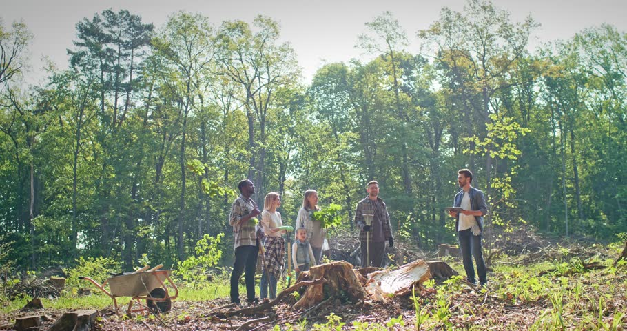 Side view of cheerful enthusiastic caring group of adults and children listening to instructions of activist planting trees in beautiful forest. Attractive multicultural people doing forestation.