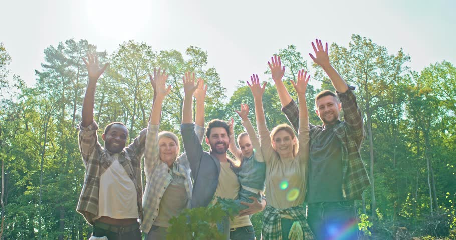 Portrait of diverse and multi-age group of enthusiastic volunteers in forest standing together and smiling with raised hands after successfully completing reforestation work. Looking at camera.
