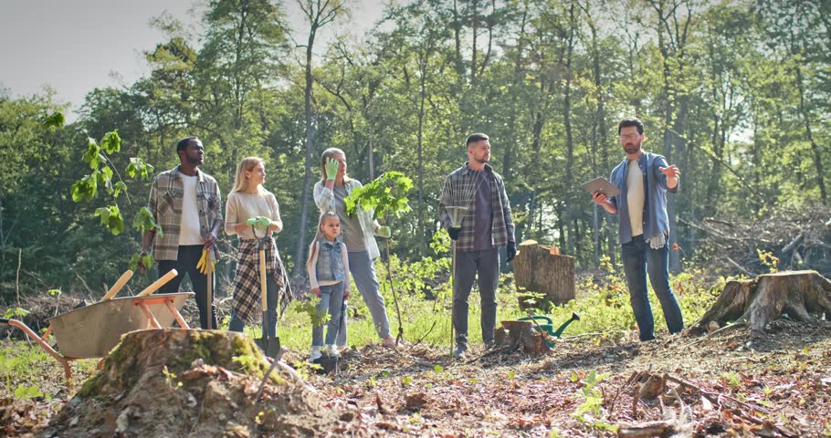 Side view of beautiful caring group of people helping nature doing forestation work listening to instructor. Enthusiastic kids and adults volunteers planting trees in nice forest. Nature concept.