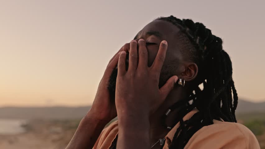 In a slow-motion, close-up shot, a black male closes his eyes, gently touches his face with his hands, and embraces a moment of self-awareness during a captivating sunset Royalty-Free Stock Footage #1109224575