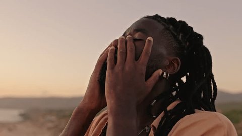 In a slow-motion, close-up shot, a black male closes his eyes, gently touches his face with his hands, and embraces a moment of self-awareness during a captivating sunset: film stockowy