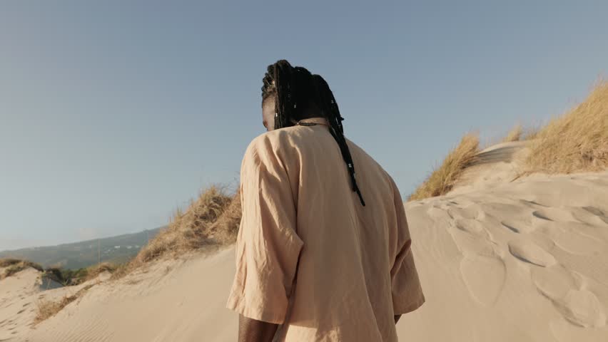 An African black male with stylish dreads and a loose open shirt, exudes confidence while turning to the camera against a sandy dunes background Royalty-Free Stock Footage #1109224577