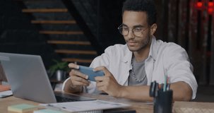 Carefree African American man playing online video game with smartphone relaxing in workplace in creative office. Millennials and entertainment concept.