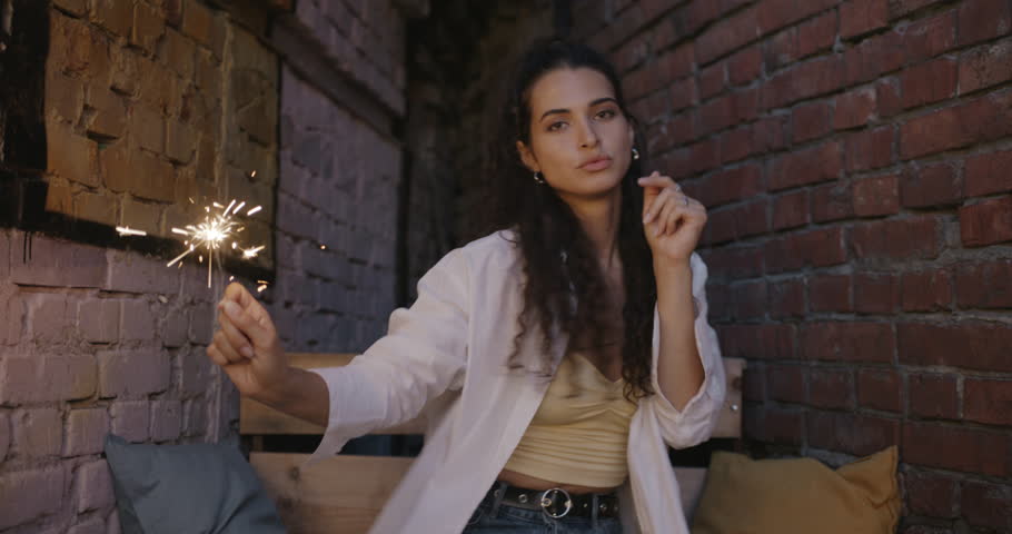 Portrait of joyful girl dancing with sparkler enjoying music sitting in open air cafe loft style in summer evening. Happiness and holiday concept. | Shutterstock HD Video #1109226685