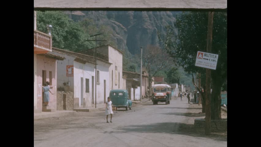 Mexico CIRCA 1960s: A town street in Mexico. A girl crosses road. A small bus drives down dirt road and stops, and people board. Women in headscarves walk.