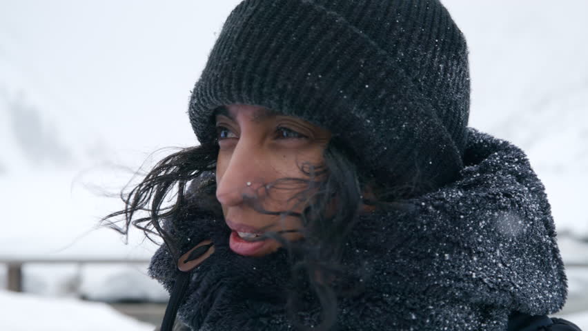 Medium close-up of Indian middle-aged woman with dark skin, nose piercing in black knitted hat looking into camera, smiling, outdoors during blizzard. High quality 4k footage Royalty-Free Stock Footage #1109235417
