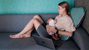 Caucasian mother combining work and childcare. Woman with child in arms works at laptop.