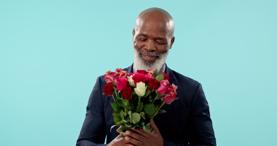 Smile, smelling flowers and a man with bouquet for valentines day on a blue background in studio. Portrait, love and romance with a happy senior person in a suit giving a rose gift of affection