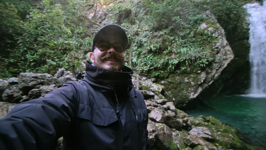 Smiling man is taking selfie against waterfall and small lake in autumn forest. Adult caucasian hipster in cap and jacket is walking in woodland and enjoying nature. Cascade view falling from rock
