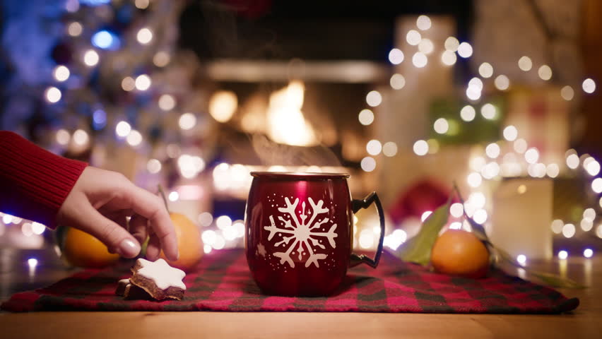 Female hand in red sweater soak in hot tea star shape gingerbread cookie on blurred christmas tree light and burning fire background. Cozy Christmas eve in festive decorated living room with fireplace