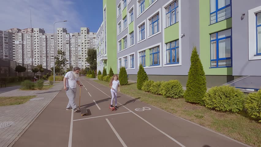Child girl teaches an old man to ride a skateboard. They are training to ride a skateboard on the street. Grandpa is riding a skateboard.