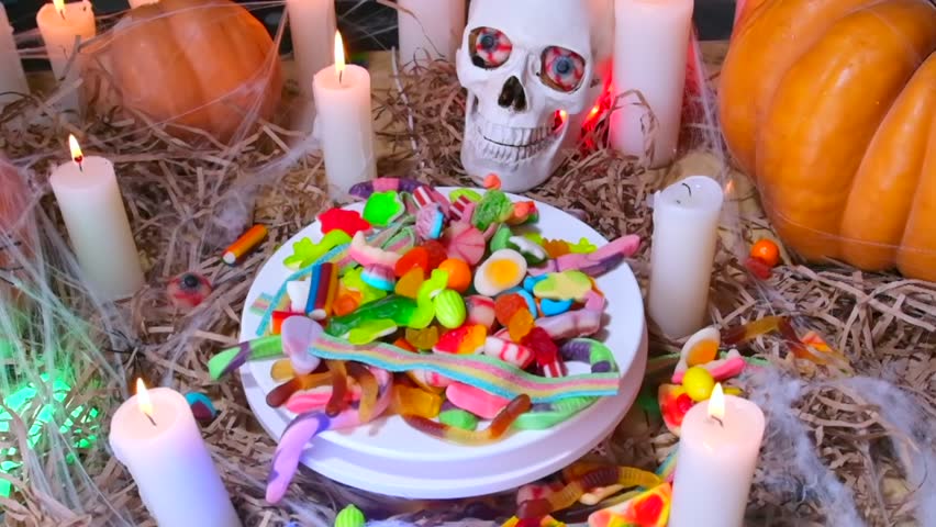 Bunch of sweets of different colors and shapes fall on plate of candy slowly rotate, of barn among candles and traditional pumpkins, close up. Frightening and unforgettable Halloween invitation.