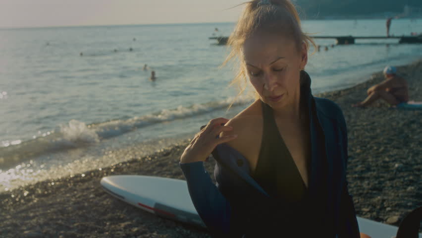 An elderly woman puts on a swimming wetsuit on the seashore. Senior female is getting ready to ride a surfboard in the sea. The old woman lives an active alternative lifestyle Royalty-Free Stock Footage #1109266761