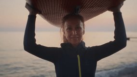 Portrait of a senior woman on the beach of the sea in a wetsuit. Granny the female is surfing on a paddle board at sunset at dusk. Active old age on the ocean. Holds the board above his head - Powered by Shutterstock - Get 15% off with code: PIKWIZARD15
