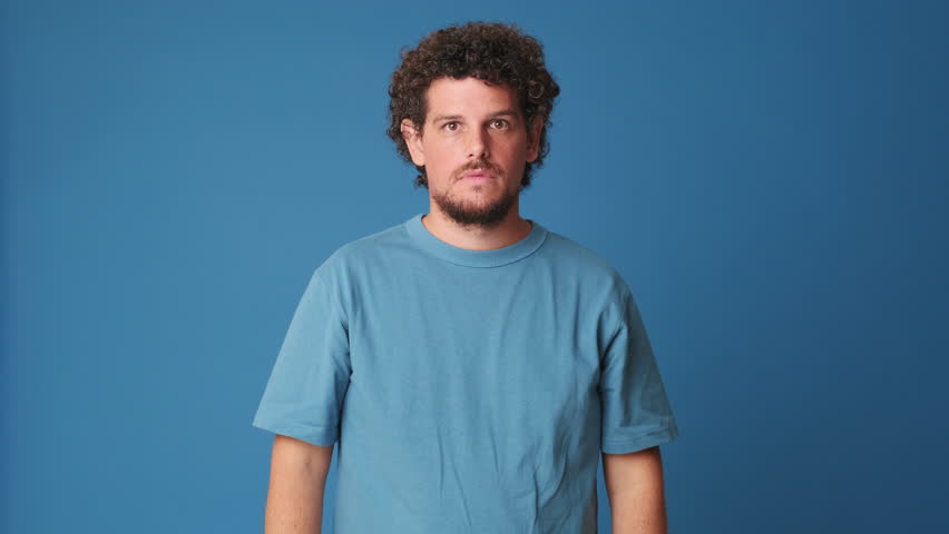 Surprised guy with curly hair dressed in blue t-shirt looking at the camera with big eyes, raises his hands says wow, delighted with the good news isolated on blue background in the studio Royalty-Free Stock Footage #1109270891