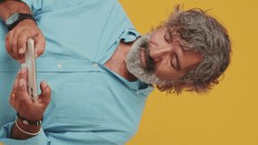 Vertical video, Elderly smiling white-haired bearded man wears a blue shirt, plays video games on a mobile phone, isolated on an orange background in the studio