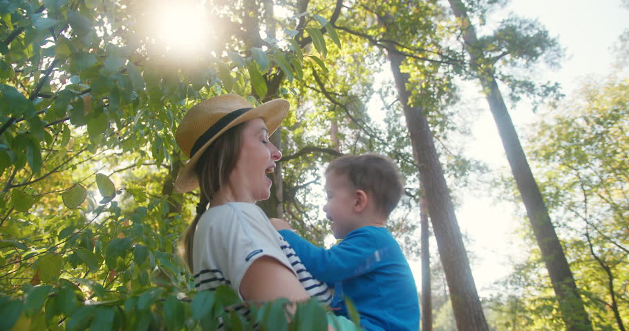 Caucasian mother and son enjoy time in nature together. The mother is holding her toddler son as they stand together in the woods. The sun is shining and they are sharing a laugh together.