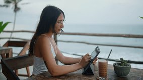 Pretty woman working remotely on tablet in a cafe by the ocean. Caucasian girl freelancer draw on device at exotic location. Young designer creating illustration sitting in tropical restaurant. - Powered by Shutterstock - Get 15% off with code: PIKWIZARD15