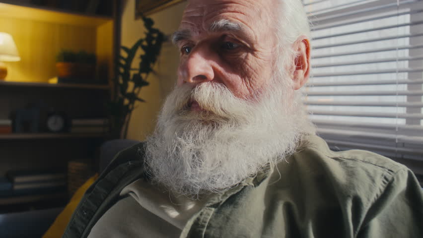 Close-up shot of face of 60-year-old retired man with grey hair, thick beard and moustache, in casual clothes sitting at home and having conversation with someone on video call Royalty-Free Stock Footage #1109276285