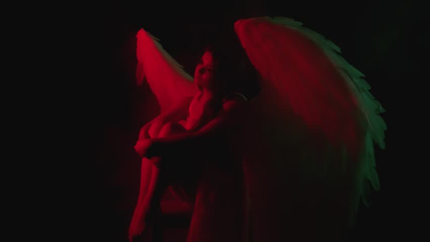 Dark silhouette Beautiful sad woman fallen angel sitting in dark hell. Costume with white wings. Black night background sky universe space. Fantasy girl goddess, fashion model posing. Red neon light