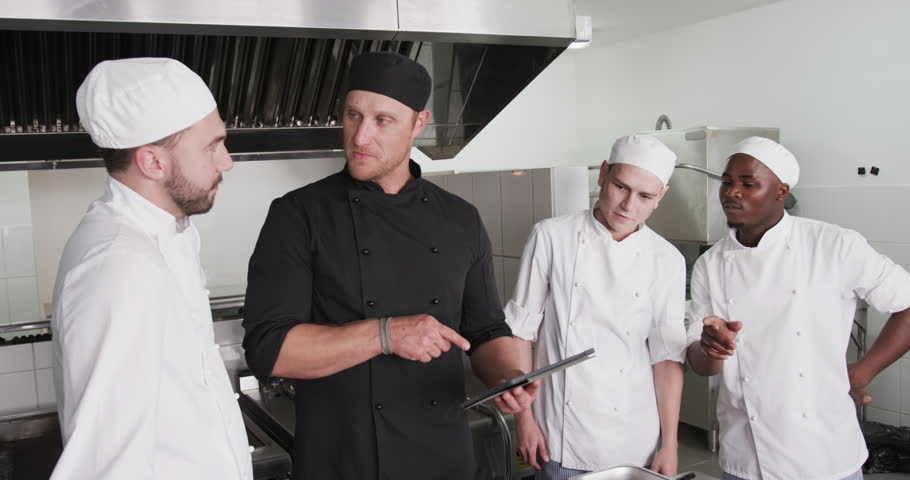 Diverse male chef instructing group of trainee male chefs using tablet in kitchen, slow motion. Technology, cooking, profession, food, training, teaching, restaurant and catering, unaltered.