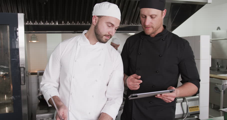 Focused cucasian male chef instructing trainee male chef using tablet in kitchen, slow motion. Technology,cooking, profession, food, training, teaching, restaurant and catering, unaltered.