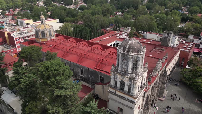 A drone flies over Coyoacán city center, Mexico City, capturing the traditional charm of the plaza and church, and the juxtaposition of the modern Torre Mitikah