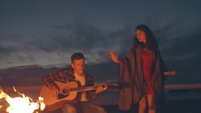 Picnic of young people man and woman with bonfire on beach in the evening. Cheerful friends singing songs playing guitar. Slowmotion shot. Time together at nature, weekend vacation. Summer activity - Powered by Shutterstock - Get 15% off with code: PIKWIZARD15