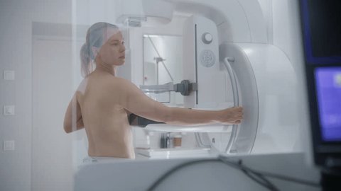 Adult woman stands topless during mammography screening checkup in hospital radiology room. Doctor presses button on control panel to launch mammogram machine using computer. Breast cancer prevention.の動画素材