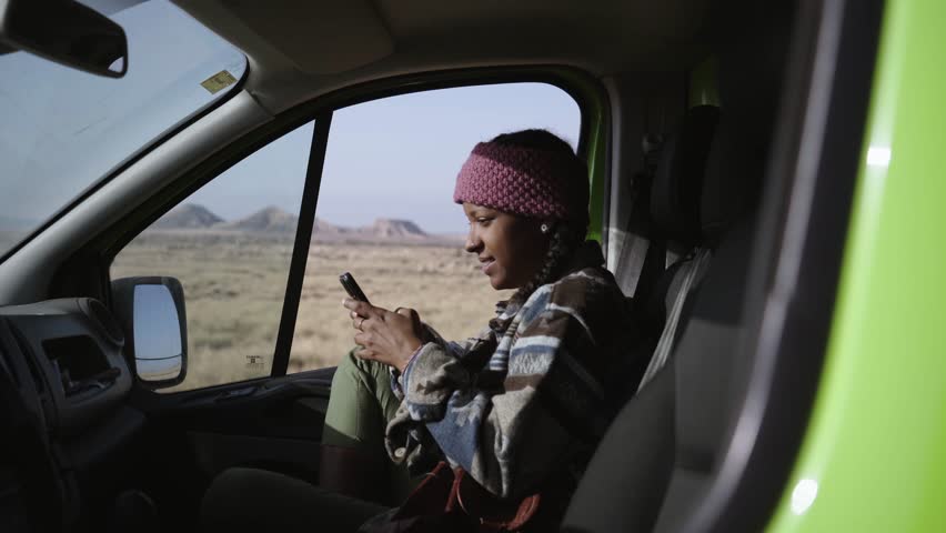 Young African woman using cell phone smiling inside camper. Black girl co-driver typing mobile laughing while resting with background of bardenas reales. Generation z road trips on nature vacation.