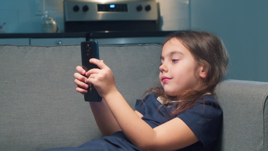 Young Girl Uses a Mobile Smartphone and Types, Plays in the Browser, Watches the Internet While Lying on the Sofa in the Kitchen. Girl Child Resting at Home Uses Smartphone for Games and Learning.