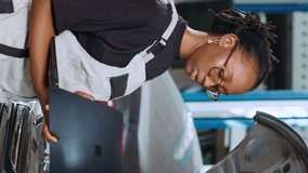 Vertical video African american mechanic in repair shop using laptop to calculate repairments invoice for client after fixing car malfunctions. Woman using device to determine final costs after