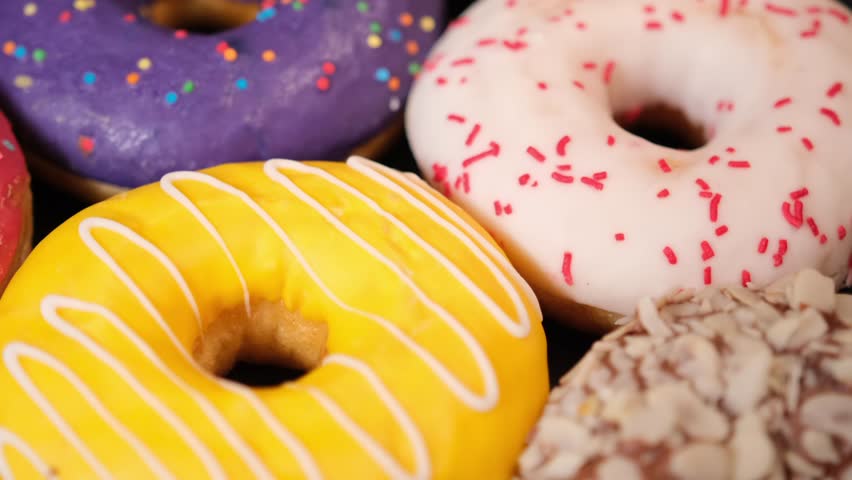 Close up colorful donuts with cream chocolate glaze and sprinkles on black background rotate. Blueberry donuts with cocoa frosting, fresh sweet buns in bakery shop display case.