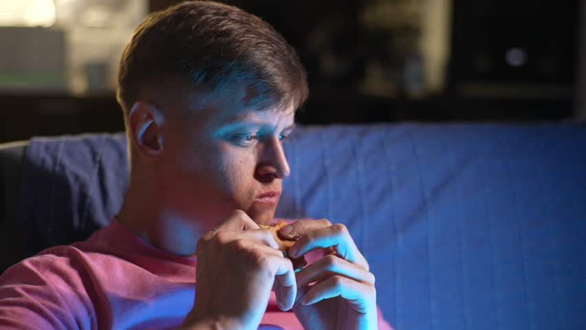Front view of relaxed young man eating tasty burger and watching movie on laptop resting on sofa in dark living room in evening at home. Happy tired male spectator enjoy online live sport match. Royalty-Free Stock Footage #1109301981