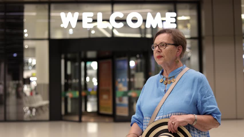 Senior woman waving at a indoor bus station with welcome sign in the back