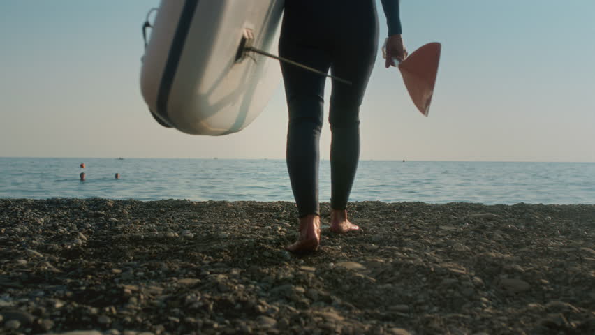 Senior woman on the beach in a wetsuit enters the water with a paddle board. An old female surfs at sunset at dusk. Active old age on the ocean. Granny steps into the sea with an oar in her hands.