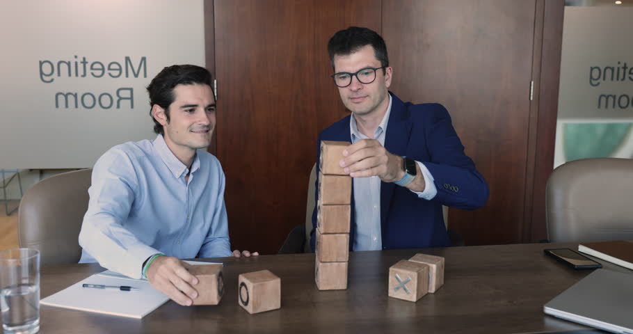 Two businessmen, multi-ethnic colleagues build tower from wooden cubes gathered in modern meeting room, take part in teamwork, motivated in partnership, strategy development, business growth, synergy | Shutterstock HD Video #1109318389