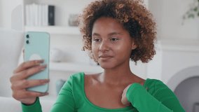 Young positive sociable African American woman waves hand in greeting making video call on smartphone and communicating with friends or relatives via internet sits on sofa in living room.