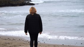 A woman walks along a sandy beach towards the ocean in slow motion.