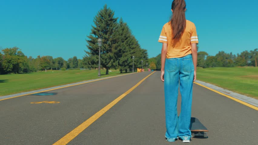 Rear view of adorable cool teenage girl skateboarder in casual outfit skateboarding on park road , enjoying freedom and active lifestyle while spending leisure in public park on summer day.