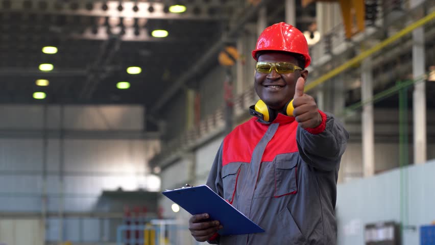 Smiling Industrial African American Worker in Personal Protective Equipment Giving Thumb Up. Portrait Of Black Industrial Worker In Red Helmet, Yellow Safety Goggles And Work Uniform In A Factory.