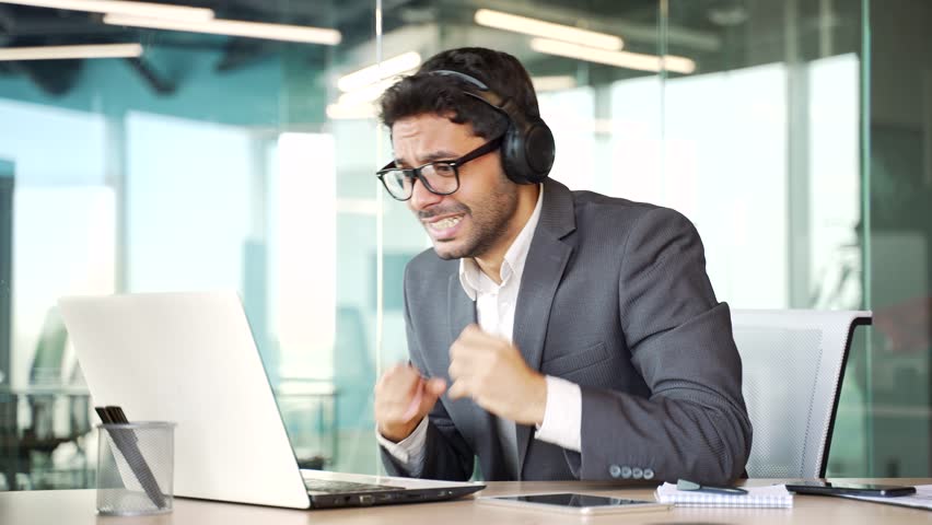 Excited young businessman in wireless headphones watching sports match competition on a laptop in business office. Happy entrepreneur in formal suit cheering for favorite team with emotional gesturing | Shutterstock HD Video #1109325291