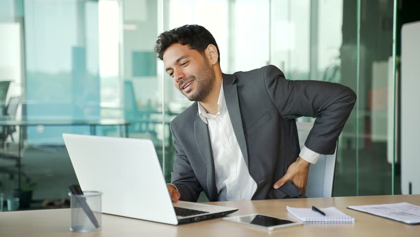 Tired young businessman in a suit suffering from back pain working on laptop sitting at workplace in business office. Sad entrepreneur massages painful lower back muscles. He is exhausted, overworked | Shutterstock HD Video #1109325295