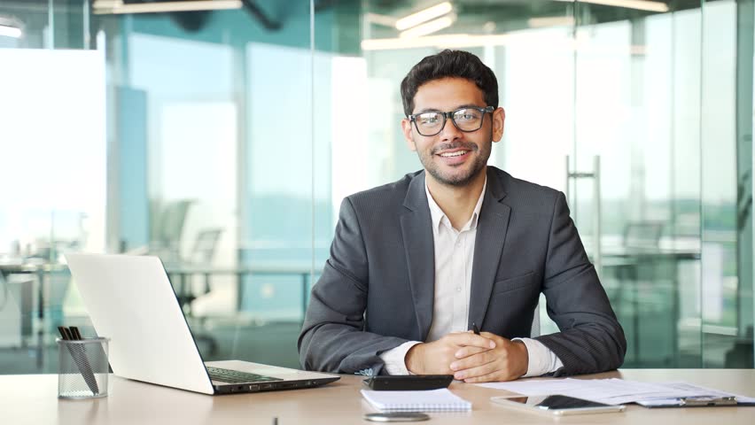 Portrait of a young successful businessman in a formal suit sitting at a workplace at a desk in a business office. Confident smiling entrepreneur in glasses looking at camera. Head shot of a manager | Shutterstock HD Video #1109325335