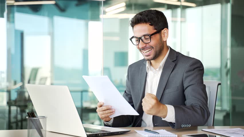 Excited financier in a formal suit is satisfied with the results of a financial report he is reviewing while sitting at a workplace in a business office. Entrepreneur is happy with positive indicators | Shutterstock HD Video #1109325351