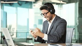 Excited young businessman in wireless headphones watching sports match competition on a tablet in business office. Happy entrepreneur in formal suit cheering for favorite team or playing computer game - Powered by Shutterstock - Get 15% off with code: PIKWIZARD15