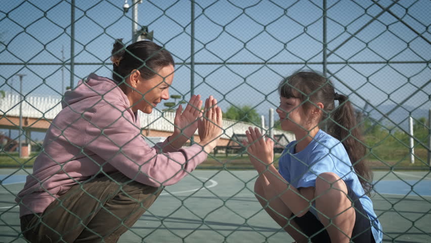 Daughter and mother squatting play with their hands on a fenced sports ground.